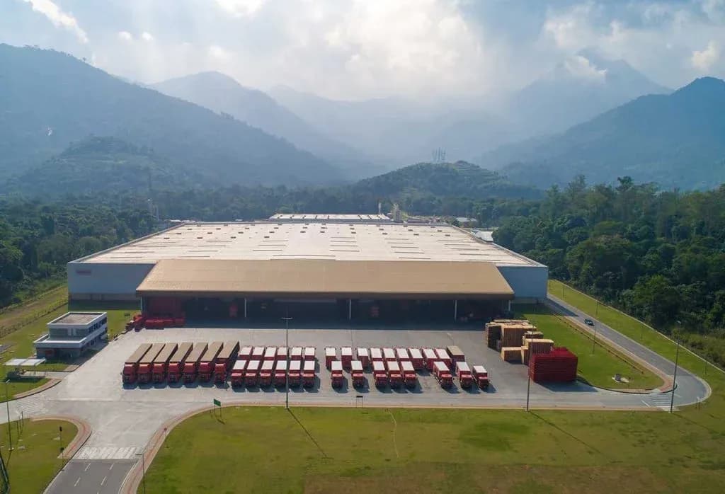 Vista aérea da fábrica da Coca-Cola Andina Brasil em Duque de Caxias, na Região Metropolitana do Rio de Janeiro, com cobertura de grande porte e frota de caminhões vermelhos alinhados em frente às docas, com montanhas ao fundo.