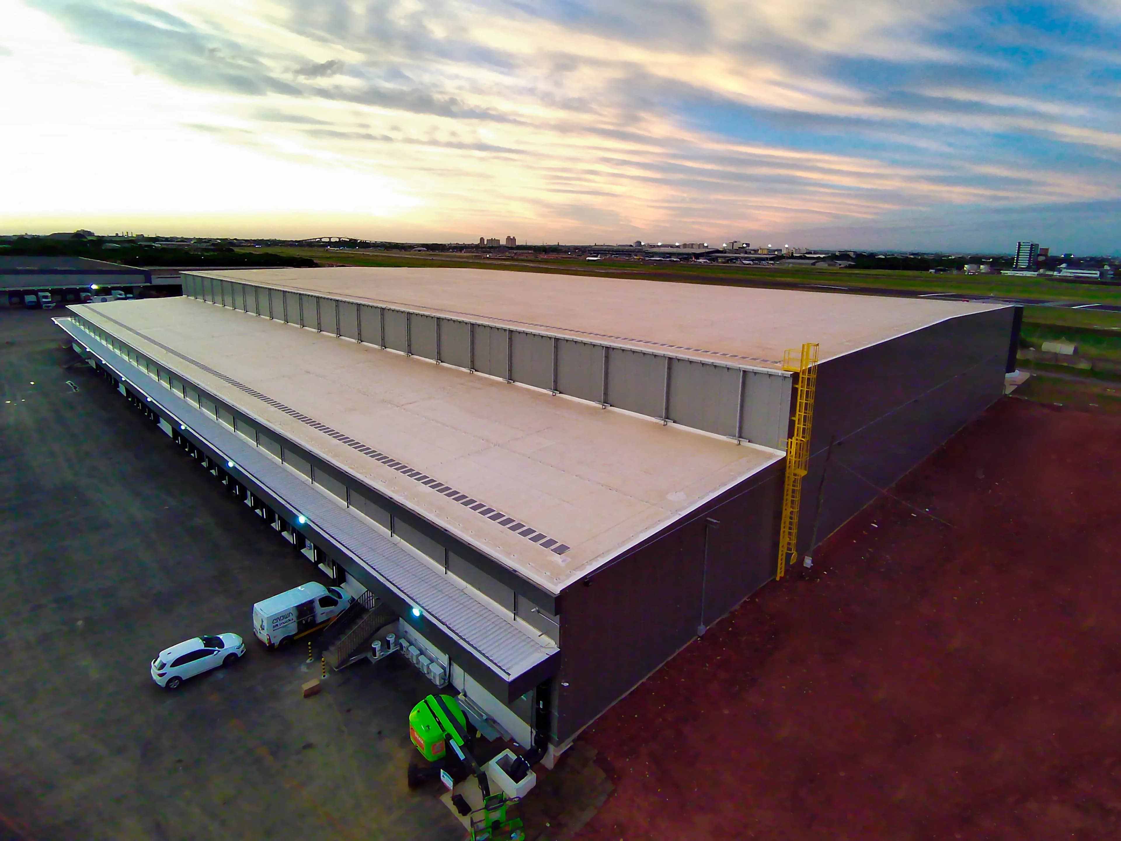 Vista aérea de galpão logístico do Grupo Zaffari ao entardecer, com cobertura bege e docas de carregamento, próximo a área aeroportuária no Rio Grande do Sul.